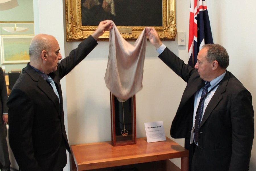 Two men lift a cloth from the Always Clock at its unveiling in Parliament House, an Australian flag beside them