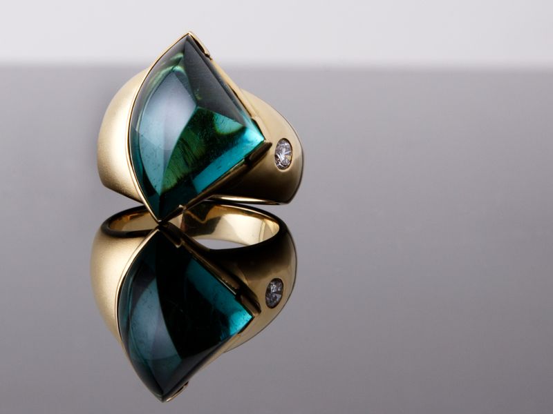 Ring with a split blue-green tourmaline cabochon rising like two sails from a yellow gold band, standing on its reflection