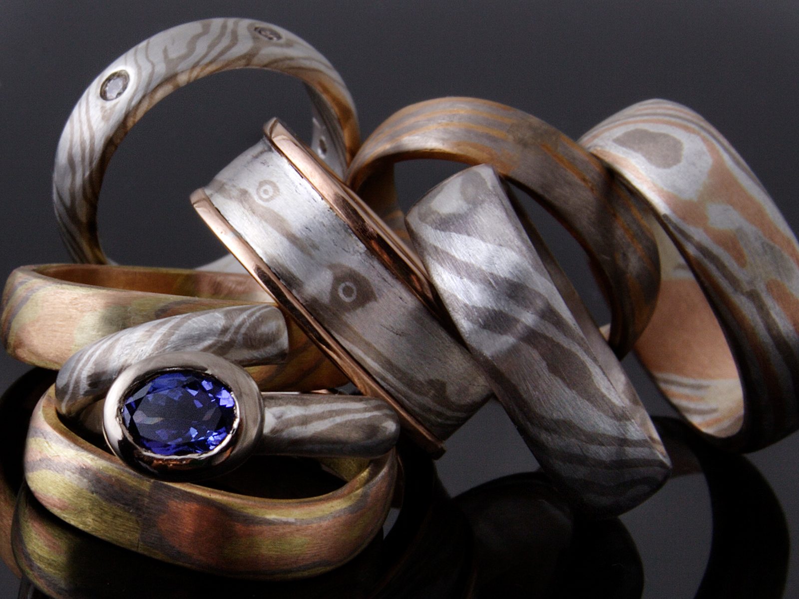 A stack of Mokume Gane rings in various combinations of metals, one set with an oval tanzanite, on black