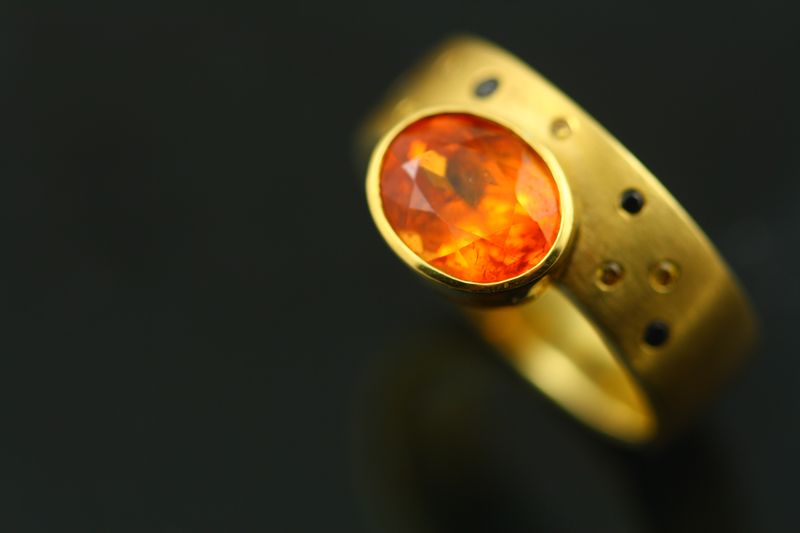 A matte yellow gold ring with an oval orange Spessartite Garnet in a full bezel, with yellow and black diamonds